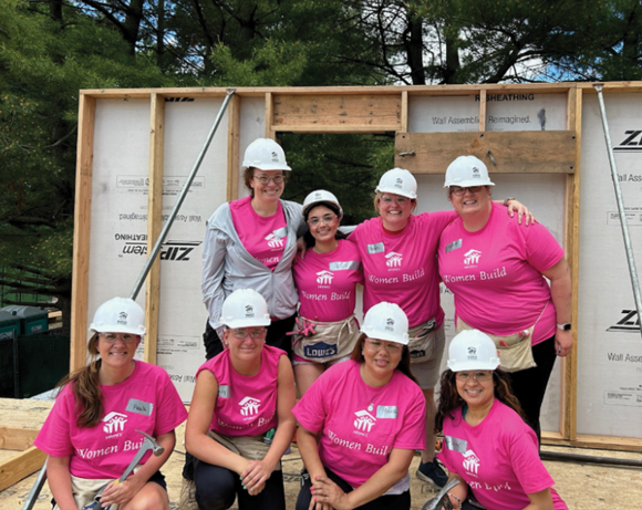 eight women in pink T-shirts and hardhats posing on a wooden construction site
